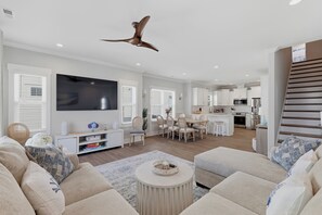 A modern living room with a flat-screen TV, a dining area with a table and chairs, and a kitchen with appliances. - Upscale New Construction 4BR/3BA Coastal Retreat • 3-Min Walk to Beach •Sleeps 9 (Carolina Beach)