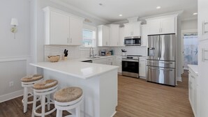 A modern kitchen with white cabinetry, a central island with stools, stainless steel appliances, and a window with blinds. - Upscale New Construction 4BR/3BA Coastal Retreat • 3-Min Walk to Beach •Sleeps 9 (Carolina Beach)