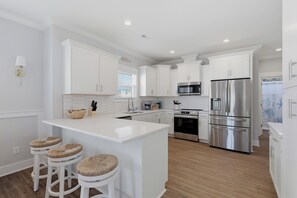 A modern kitchen with white cabinetry, a central island with stools, stainless steel appliances, and a window with blinds. - Upscale New Construction 4BR/3BA Coastal Retreat • 3-Min Walk to Beach •Sleeps 9 (Carolina Beach)