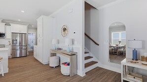 A modern kitchen with white cabinetry and stainless steel appliances, a wooden dining table with chairs, and a staircase leading to a dining area. - Upscale New Construction 4BR/3BA Coastal Retreat • 3-Min Walk to Beach •Sleeps 9 (Carolina Beach)