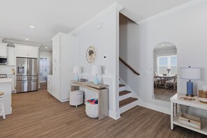 A modern kitchen with white cabinetry and stainless steel appliances, a wooden dining table with chairs, and a staircase leading to a dining area. - Upscale New Construction 4BR/3BA Coastal Retreat • 3-Min Walk to Beach •Sleeps 9 (Carolina Beach)