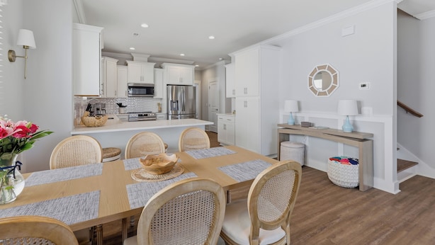 A modern kitchen with white cabinetry, stainless steel appliances, and a dining area with a wooden table and wicker chairs. - Upscale New Construction 4BR/3BA Coastal Retreat • 3-Min Walk to Beach •Sleeps 9 (Carolina Beach)