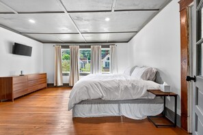 A bedroom with a bed, a wooden dresser, a TV mounted on the wall, and a window with curtains.