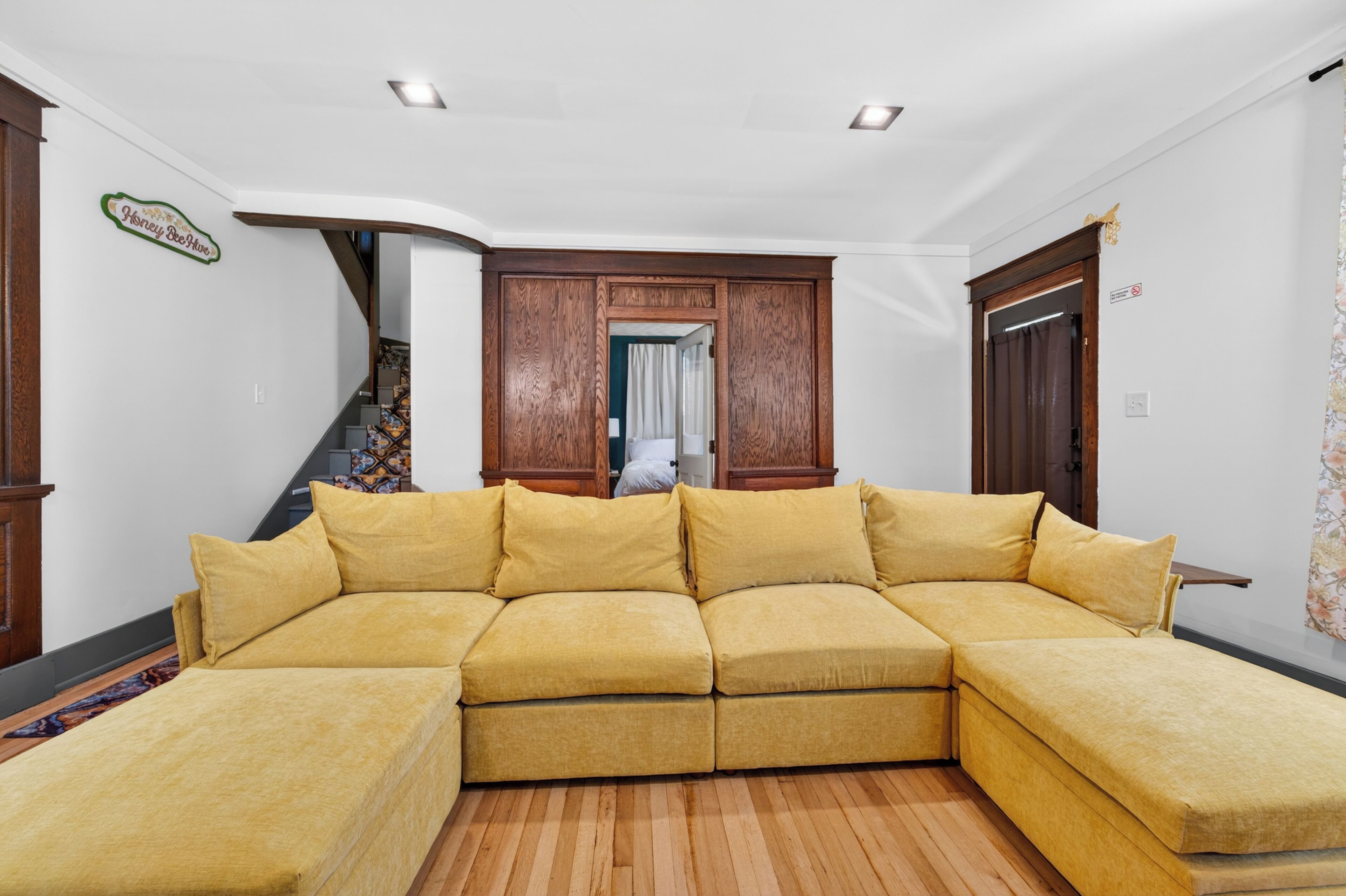 A living room with a sectional sofa, wooden flooring, a staircase, and a door with a sign.