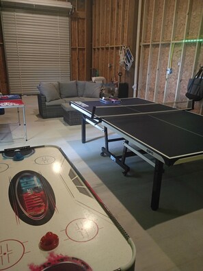 Taula de ping-pong amb un suport de pilotes i una taula de tennis en una habitació amb un sofà i una finestra tancada.
