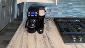 A kitchen with a coffee maker, a cabinet with mugs, and a tiled backsplash. - House, 2 Masters, Hot Tub, Tiki & Game, Sleeps 12 (Bolivar Peninsula)