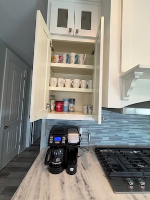 Eine Küche mit Kaffeemaschine, einem Schrank mit Tassen und einer gefliesten Spritzschutzwand.