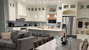 A modern kitchen with white cabinets, a stainless steel refrigerator, and a dining area with a wooden table and chairs. - House, 2 Masters, Hot Tub, Tiki & Game, Sleeps 12 (Bolivar Peninsula)
