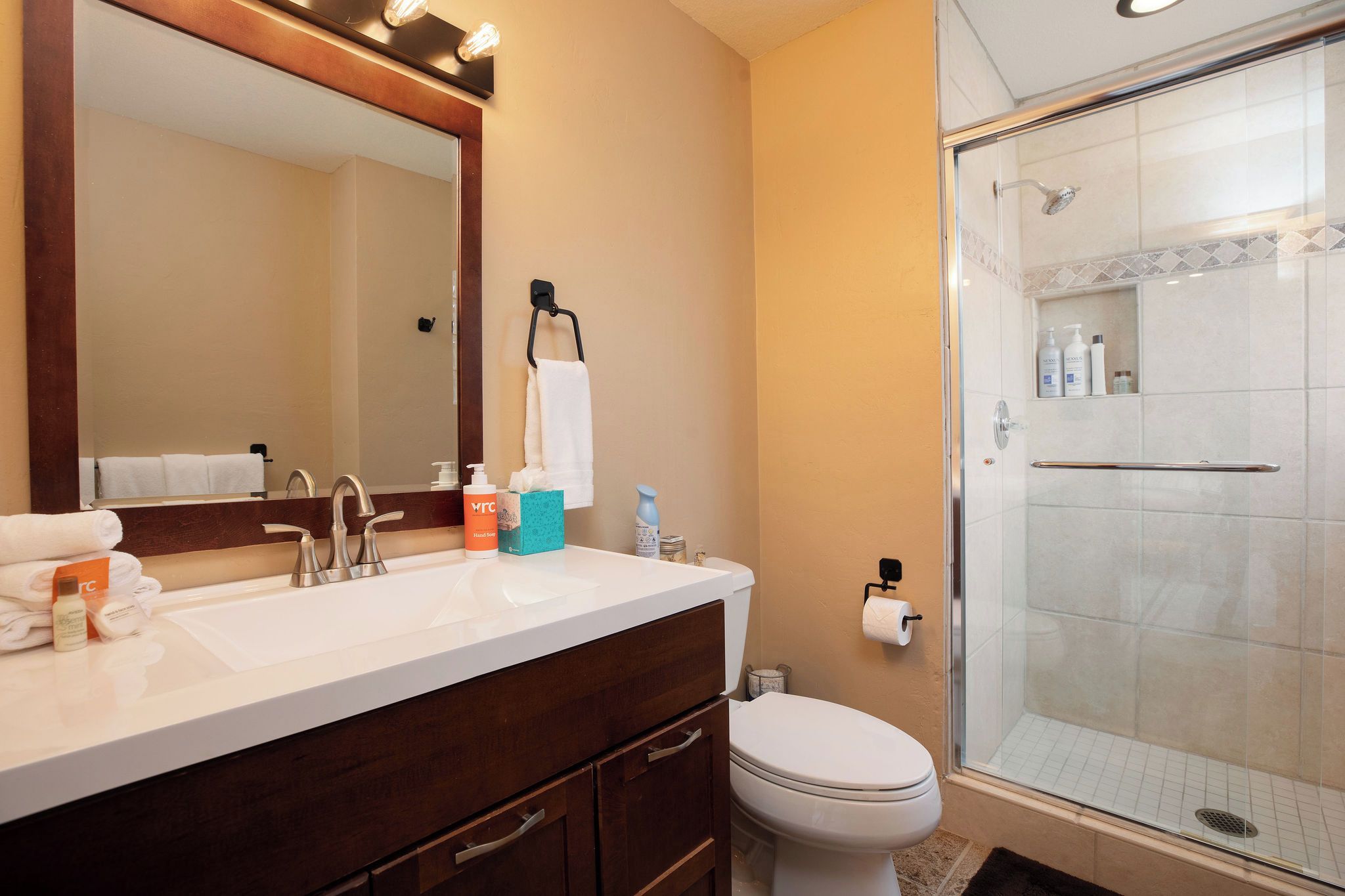 Un baño con lavamanos doble, inodoro, cabina de ducha con mampara de vidrio y espejo con iluminación superior.
