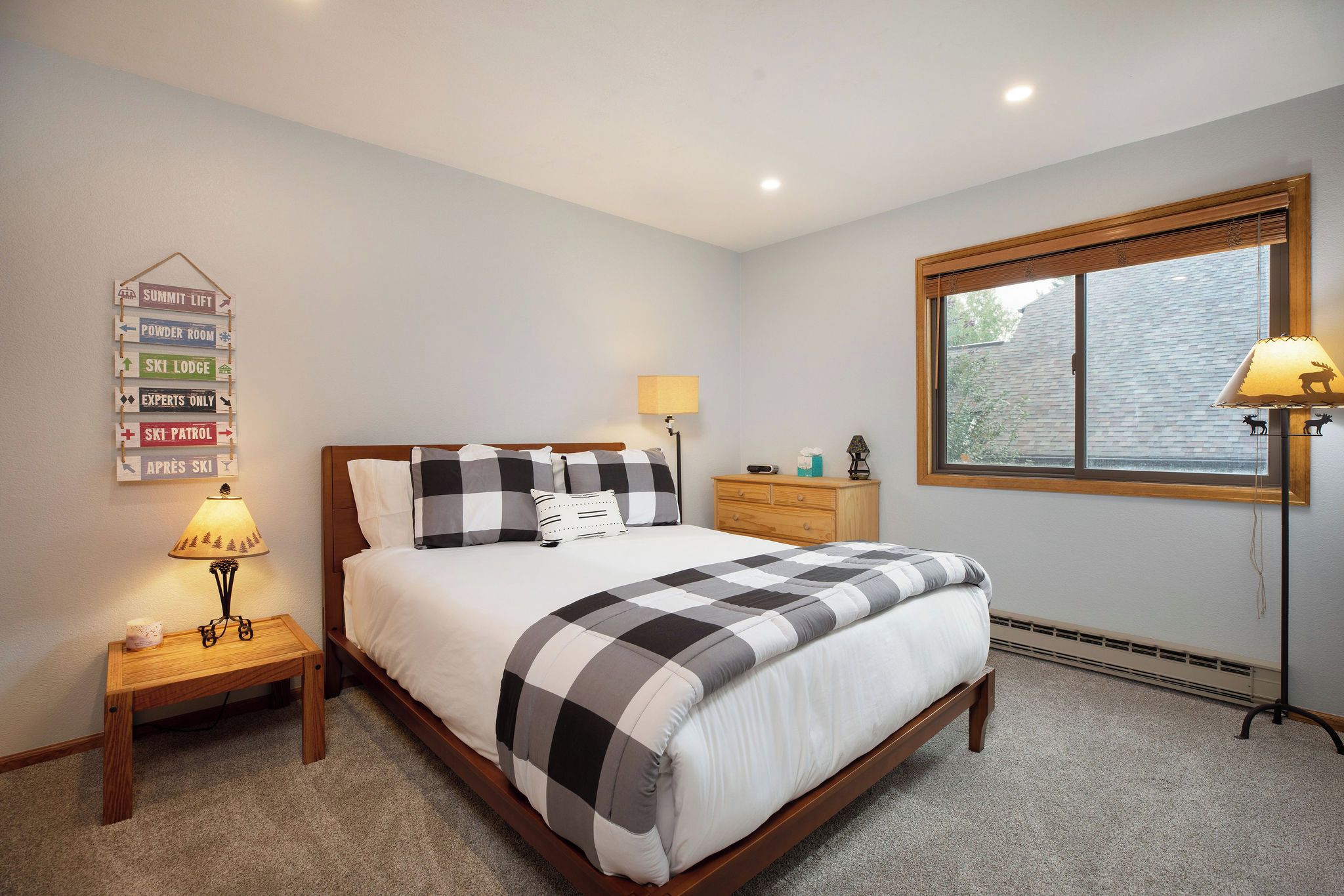 A bedroom with a bed, bedside tables, a lamp, a dresser, a window with blinds, and a wall sign.