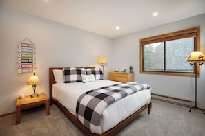 Un dormitorio con cama, mesitas de noche, lámpara, cómoda, ventana con persianas y un cartel en la pared.
