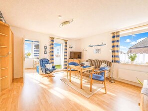 Ein geräumiges Wohnzimmer mit Holzboden, einer Couch, einem Esstisch mit Stühlen, einem Fernseher und einem Fenster mit Vorhängen.