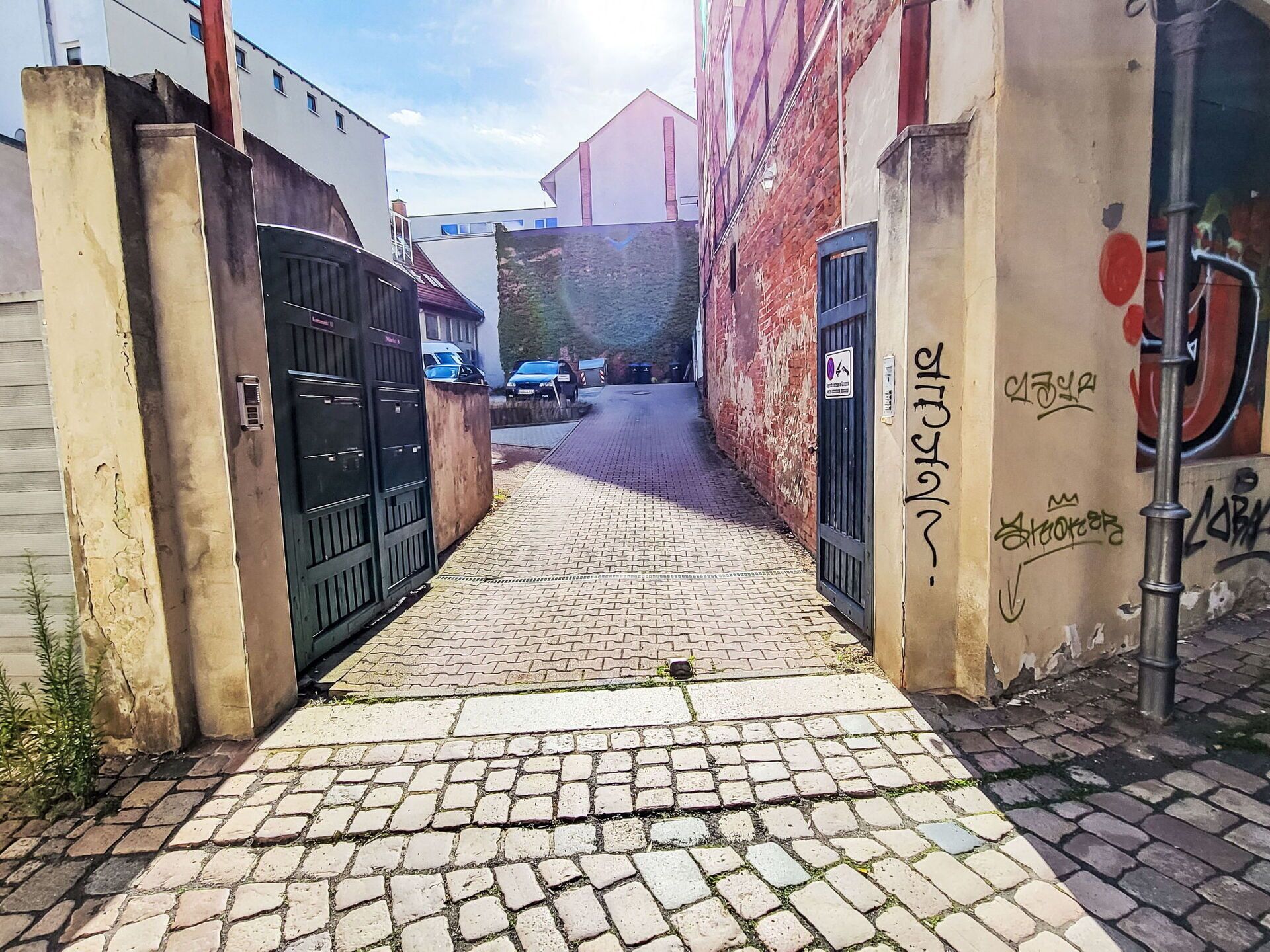 A narrow cobblestone alleyway with a black gate on the left and a wall with graffiti on the right.