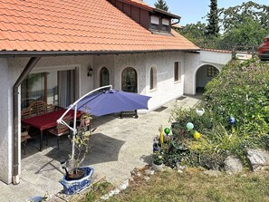 A house with a tiled roof, a covered patio with a table and chairs, and a garden with various plants and decorations.