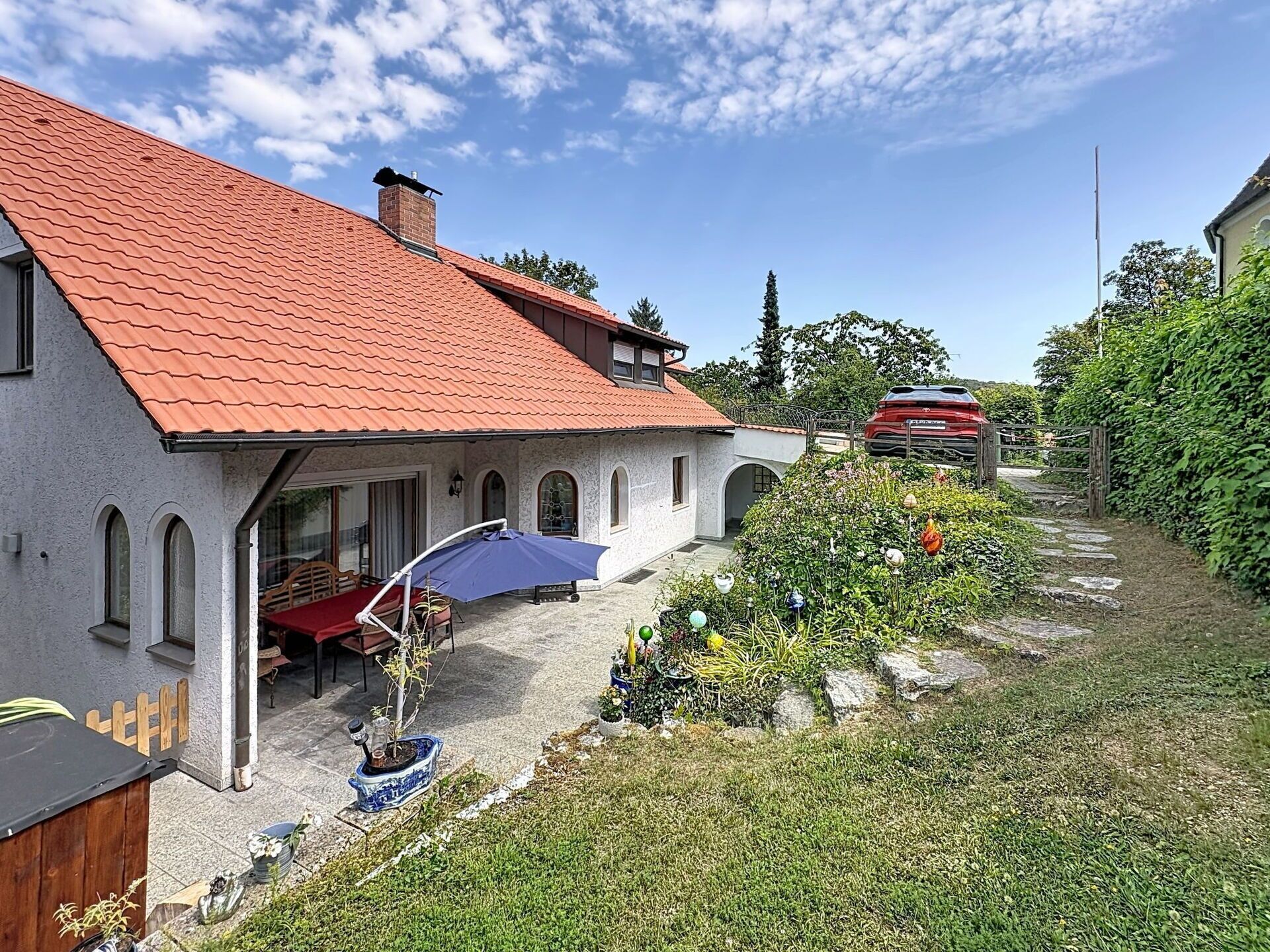 A house with a red tiled roof, a covered patio with a table and chairs, and a garden with various plants and a pathway.
