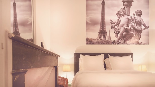 Habitación de hotel con cama, mesita de noche, lámpara y una pared con una imagen de la Torre Eiffel y una estatua.