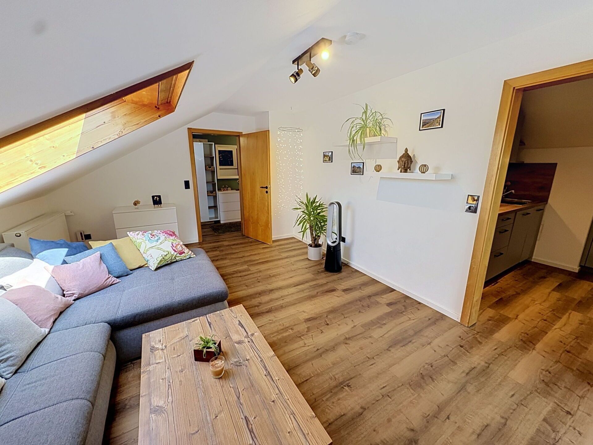 Un espacio habitable moderno estilo loft, con techo inclinado, un sofá gris, una mesa de centro de madera y una cocina visible a través de un vano abierto.
