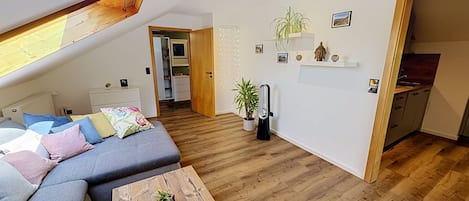 Un espace de vie moderne de style loft, avec un plafond en pente, un canapé gris, une table basse en bois et un coin cuisine visible à travers une porte ouverte.
