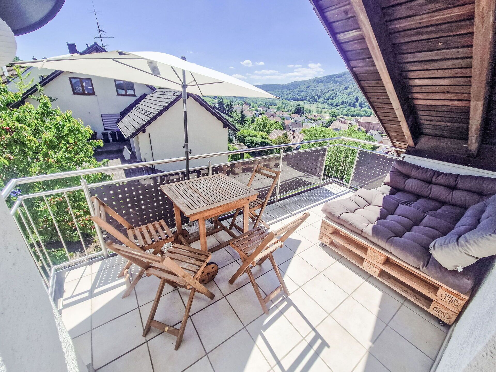 A balcony with a wooden table and chairs, a cushioned sofa, and a view of a house and greenery.