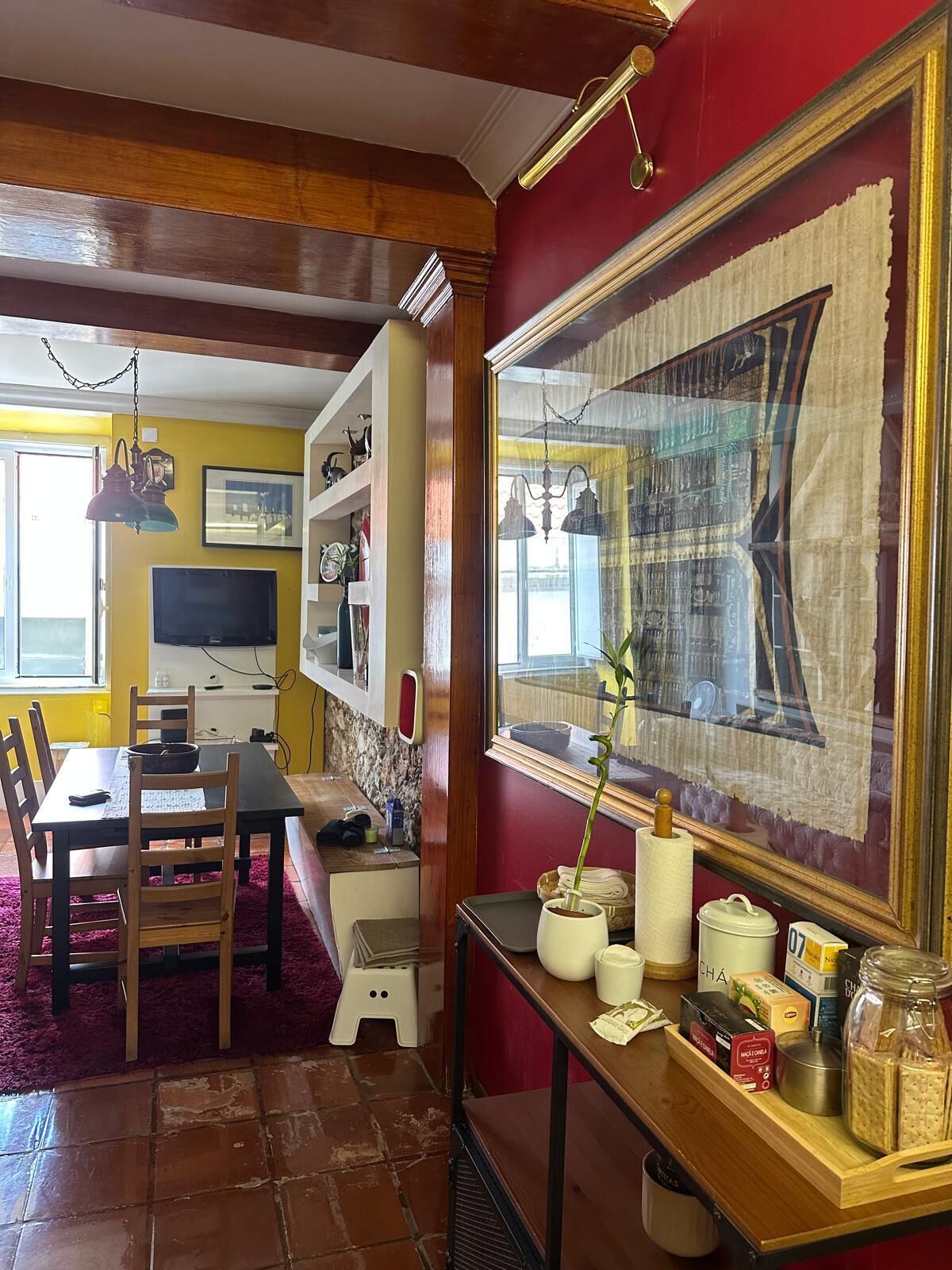 A dining area with a wooden table, chairs, and a red carpet. There is a window with a view of the outside, a wall-mounted TV, and a shelf with decorative items.