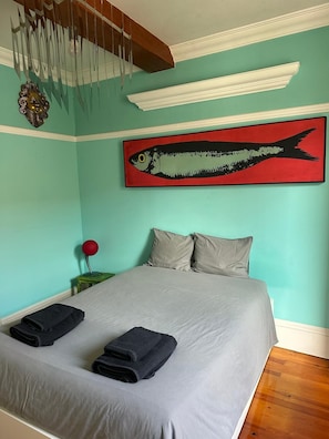 Ein Schlafzimmer mit einem Bett, Kissen, einem Gemälde von einem roten Fisch, einer Wandleuchte und einem kleinen Tisch mit einer roten Lampe.