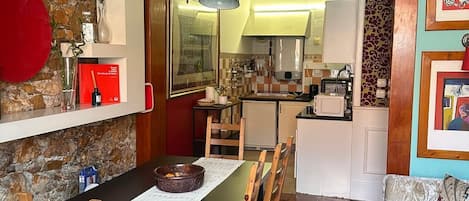 A dining area with a wooden table, chairs, and a red rug. There is a kitchen in the background with a refrigerator and a microwave.