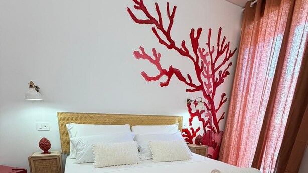 A neatly made bed with white linens and rolled towels, a red coral wall art, and a wooden headboard. - Refined structure in Milazzo, designed to experience an exclusive stay. (Milazzo)