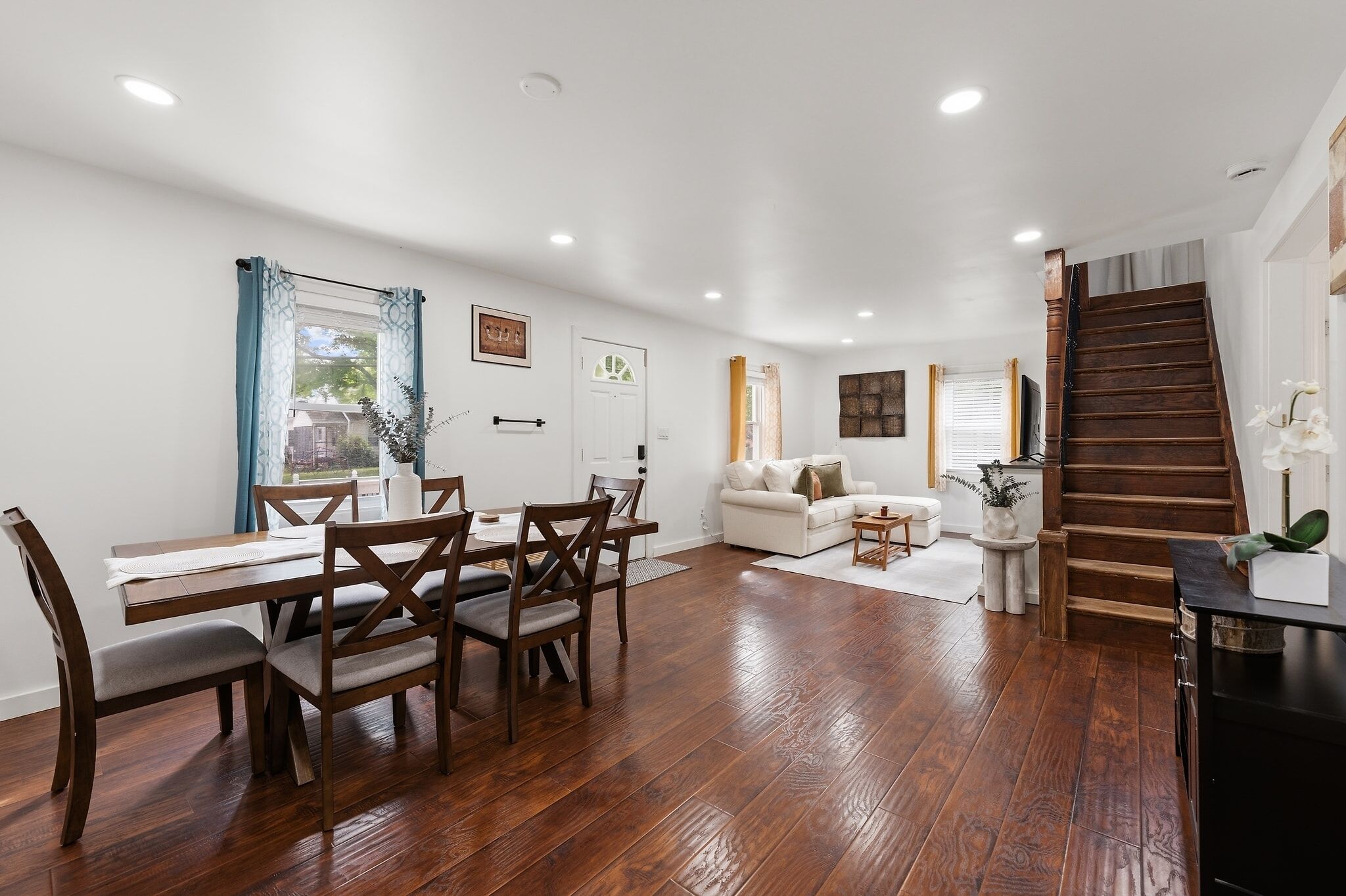 Amplia sala de estar con mesa y sillas de comedor, sofá, escalera y vistas al exterior.