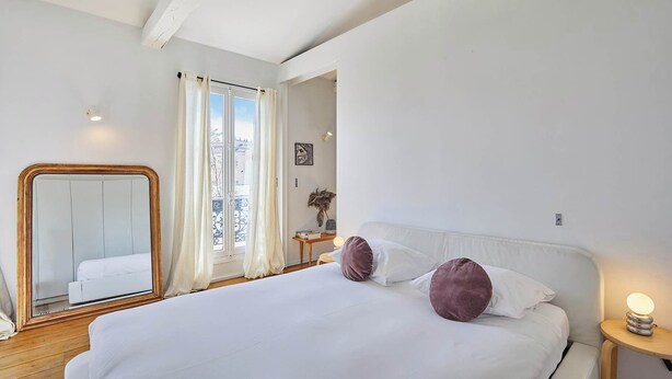 Un dormitorio con una cama grande, un espejo, una mesita de noche con lámpara y una ventana con cortinas.
