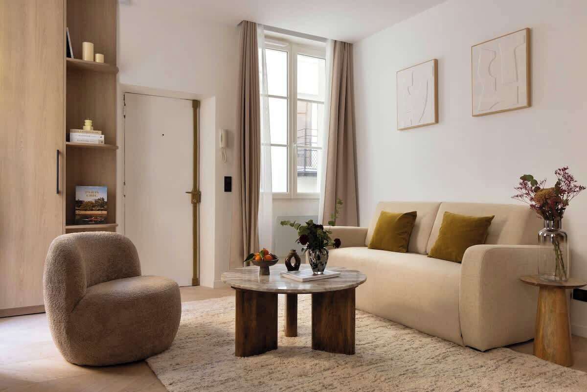 Un salon avec un canapé beige, une table basse ronde, un fauteuil beige, une bibliothèque et une fenêtre avec des rideaux.