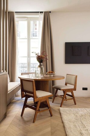 Ein modernes Wohnzimmer mit einem runden Holztisch, zwei Holzstühlen, einem beigen Sofa und einem großen Fenster mit Vorhängen.