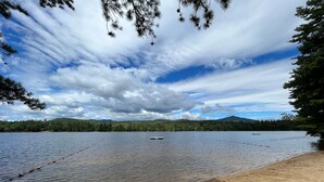 Vista para um lago com uma praia arenosa, águas calmas e uma colina arborizada ao fundo.