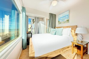 Ein Hotelzimmer mit einem großen Bett, einem Holzkopfteil, einem Nachttisch und Blick auf den Strand durch das Fenster.