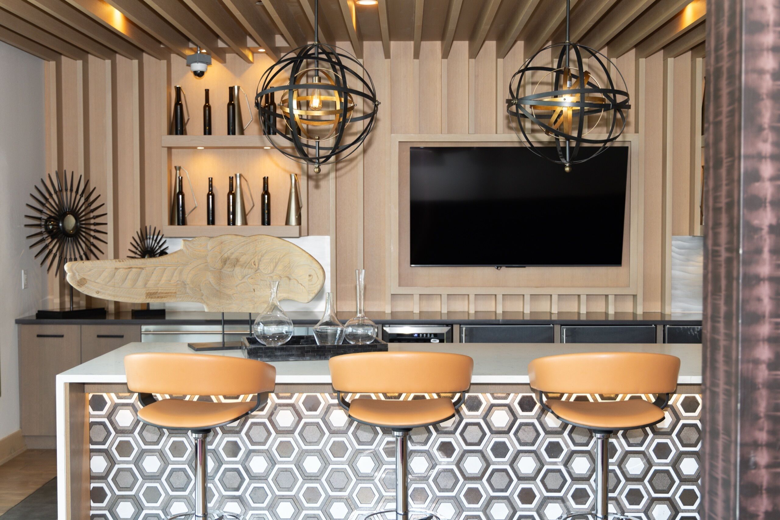 A modern kitchen with a central island, bar stools, pendant lights, and a flat-screen TV mounted on the wall.