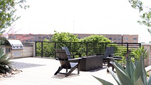 An outdoor patio with two Adirondack chairs and a fire pit, surrounded by a metal fence and greenery. - Modern Deep Ellum Loft with Rooftop Pool & Downtown Dallas Skyline Views (Dallas)