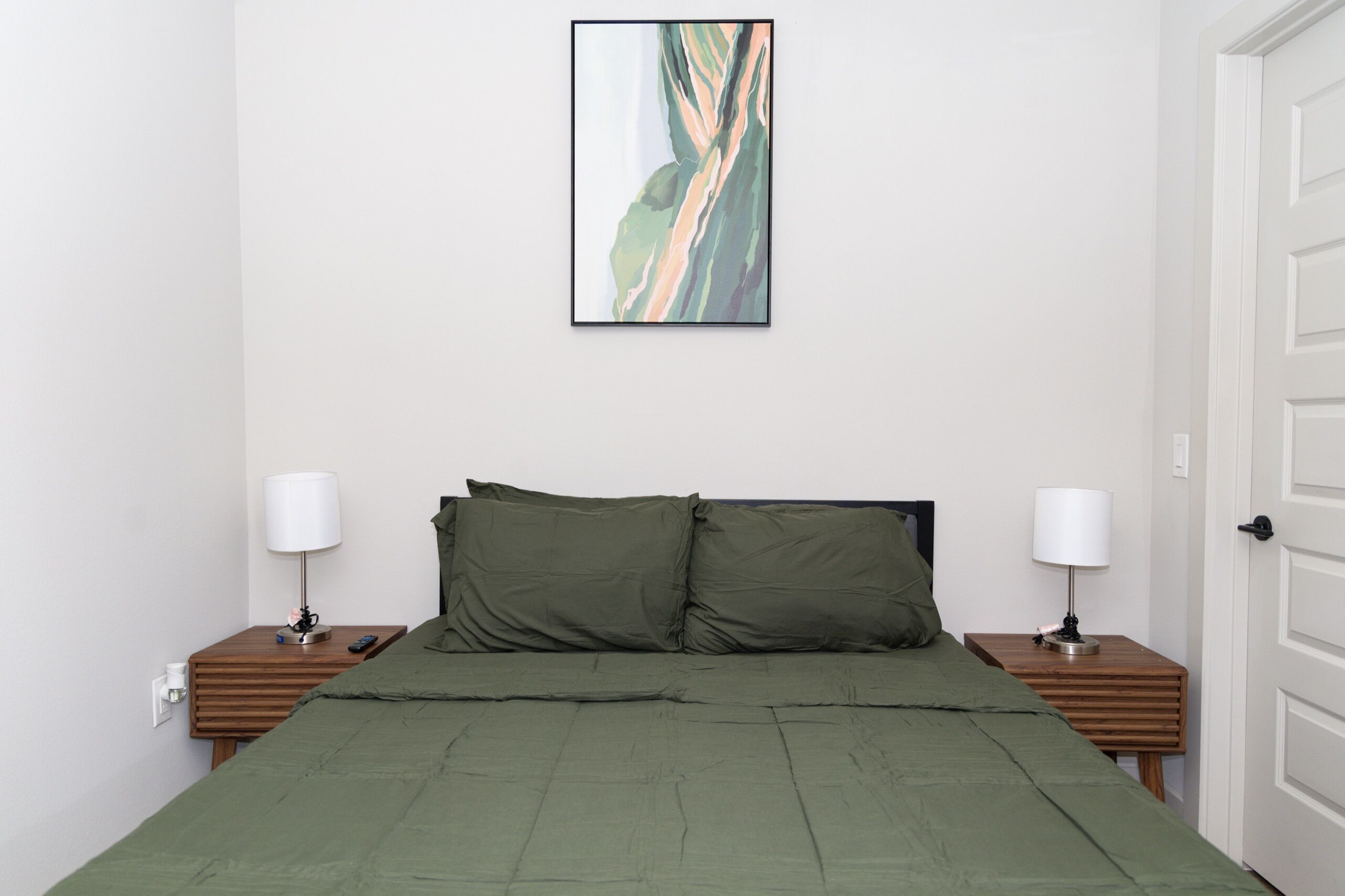 A bedroom with a bed, two bedside tables, two lamps, a door, and a framed abstract painting on the wall.
