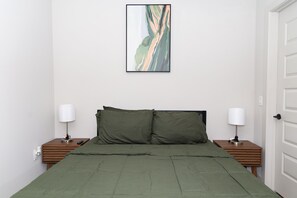 A bedroom with a bed, two bedside tables, two lamps, a door, and a framed abstract painting on the wall.
