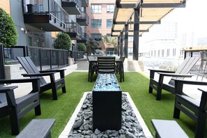 An outdoor seating area with a long table and chairs, surrounded by a low fence and greenery.