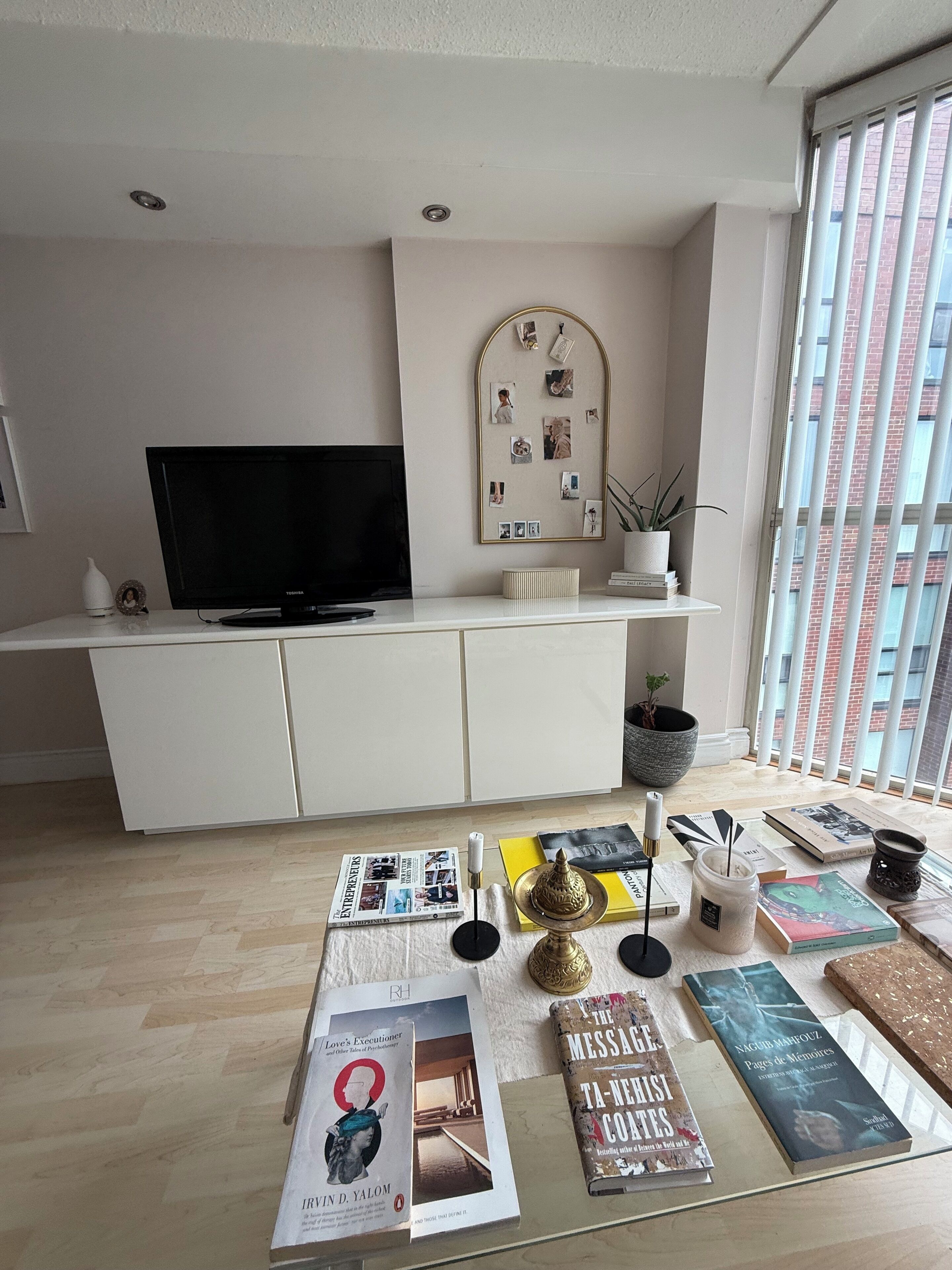 A modern living room with a flat-screen TV, a white cabinet, a wall-mounted mirror, and a window with blinds.