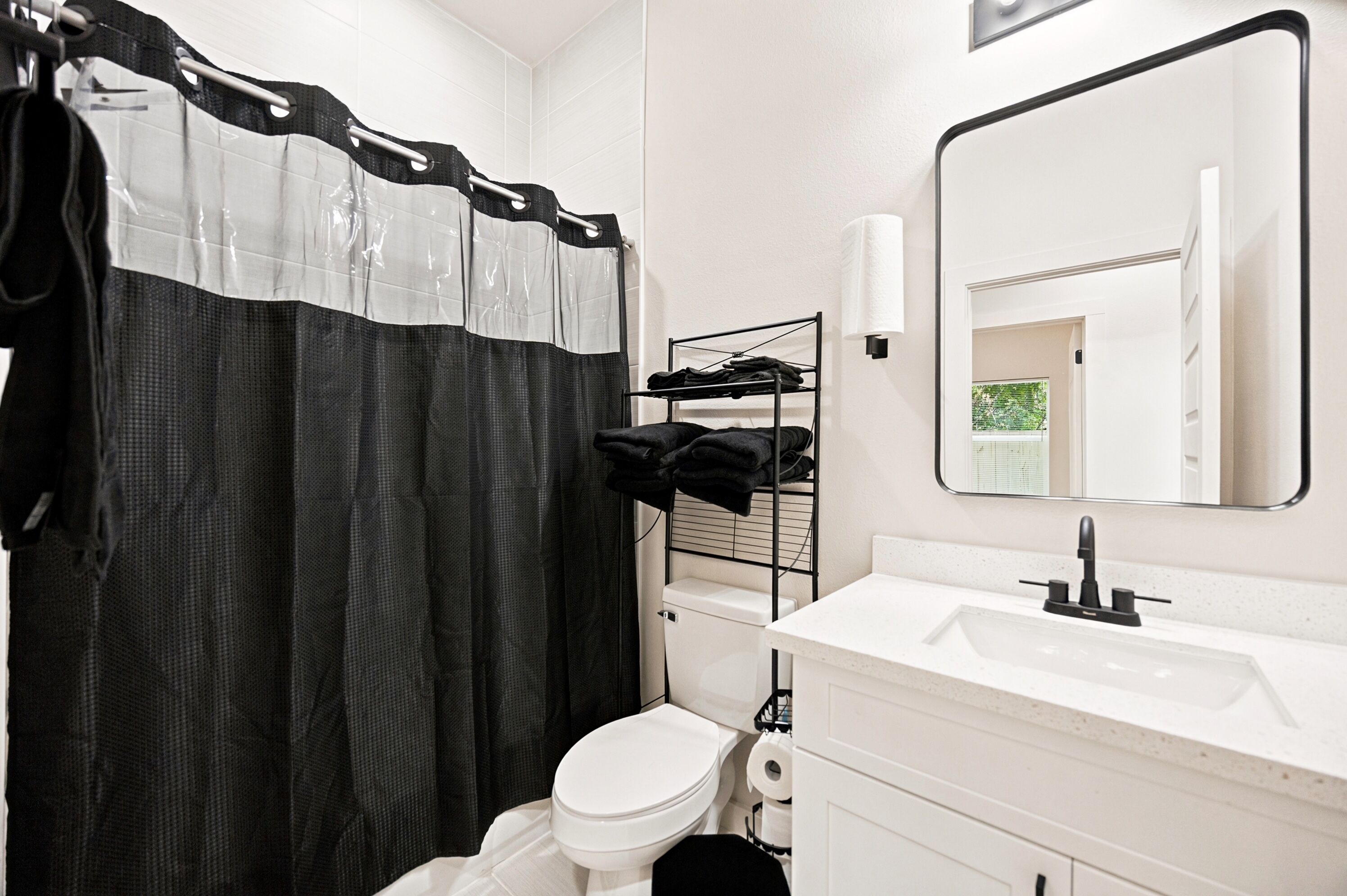 Un baño con un inodoro blanco, una cortina de ducha blanco y negro, un lavamanos blanco con un espejo y un perchero con toallas.