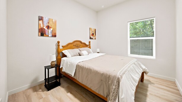 A bedroom with a wooden bed, a nightstand, two wall-mounted paintings, a window with blinds, and a wooden floor. - 3BR Modern Home, 15 Min to Downtown, Hobby, & NRG (Houston)