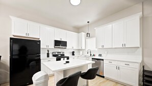 A modern kitchen with white cabinetry, a black refrigerator, a microwave, a stove, and two bar stools. - 3BR Modern Home, 15 Min to Downtown, Hobby, & NRG (Houston)