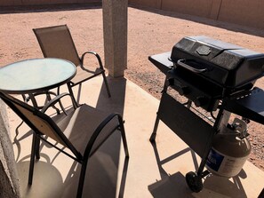 Eine Terrasse im Freien mit einem runden Tisch, zwei Stühlen, einem Grill und einer Propangasflasche.