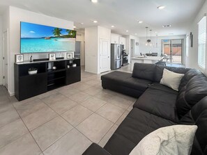 A modern living room with a large flat-screen TV, a black sofa, and a kitchen area in the background. - Modern 3BR Casa Grande Retreat | Pool Access + I-10 (Casa Grande)