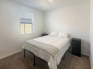 A bedroom with a bed, a nightstand, a bedside table, a window with blinds, and a carpeted floor. - Modern 3BR Casa Grande Retreat | Pool Access + I-10 (Casa Grande)