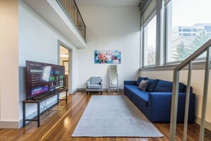 Un salon avec un téléviseur TCL Roku, un canapé bleu, un fauteuil gris, un grand tableau abstrait et une vue sur un bâtiment à l’extérieur.