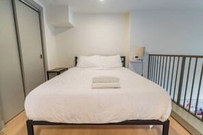 Un lit simple avec une couette blanche et une serviette pliée posée dessus, une table de chevet avec une lampe, et une vue sur le balcon extérieur.