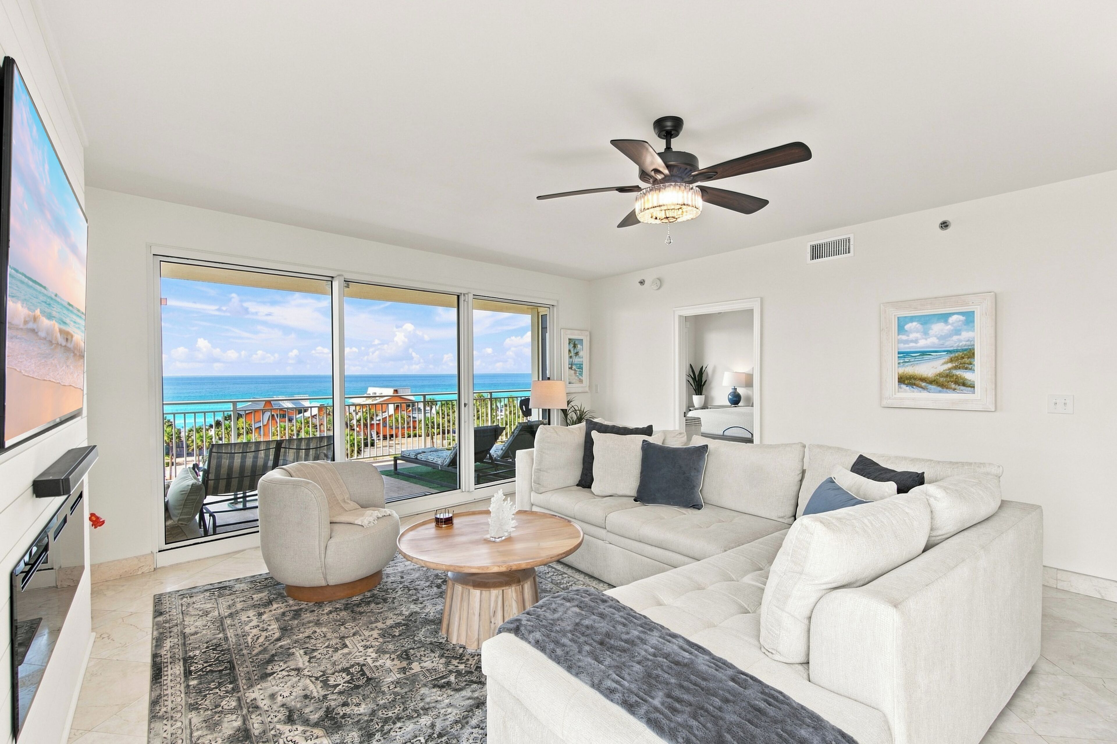 A spacious living room with a large window offering a view of the ocean, a ceiling fan, and a flat-screen TV mounted on the wall.