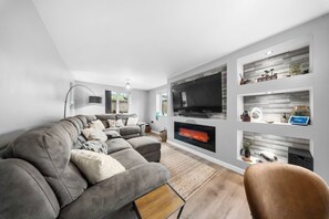 A modern living room with a sectional sofa, a flat-screen TV, a fireplace, and built-in shelving with decorative items. - Luxury Countryside Retreat with HotTub (Mildenhall)