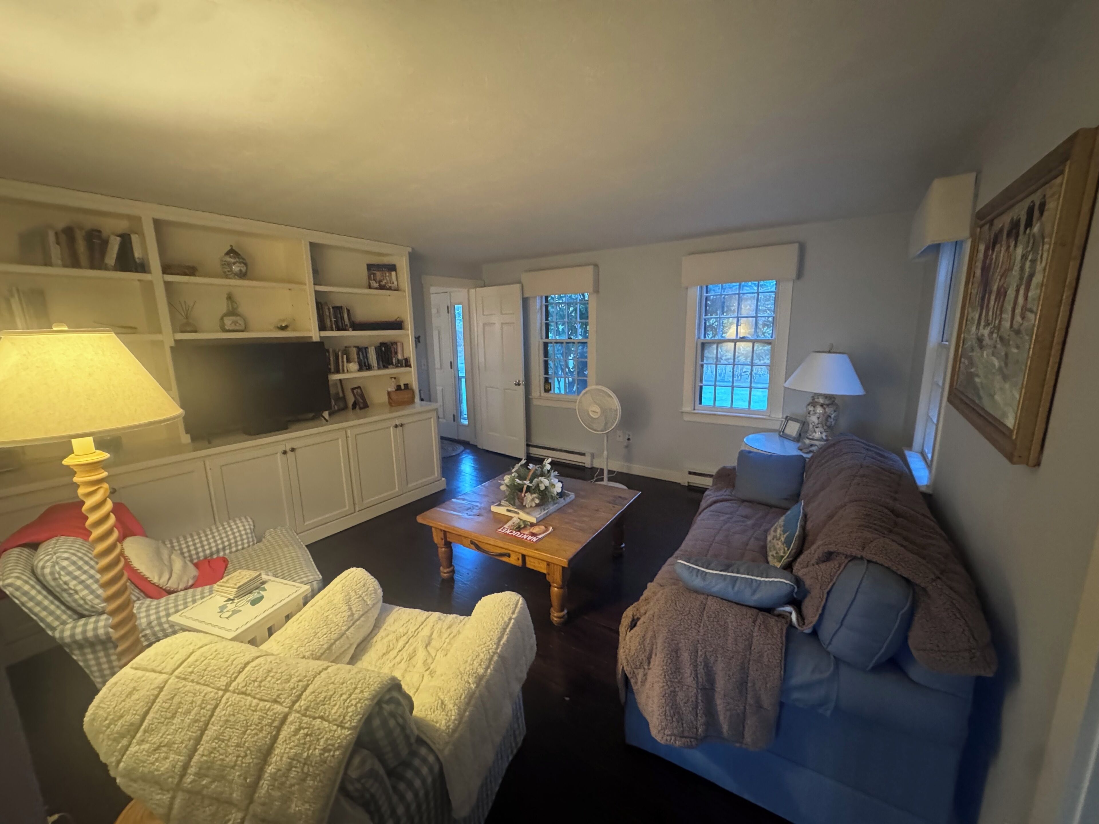 Ein Wohnzimmer mit einer Couch, Sesseln, einem Couchtisch und einem Fernseher. Regale mit Büchern und Dekoration, und eine Tür zu einem anderen Raum.